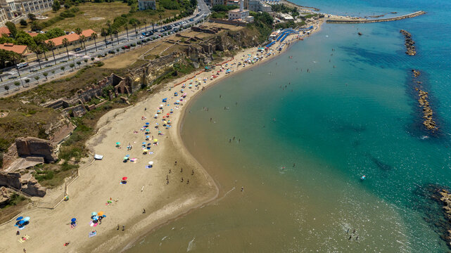Aerial view of a sunny Mediterranean beach with sandy shorelines, ancient ruins along the coast, clear turquoise sea, breakwaters, and a nearby road. Panorama of Grotte di Nerone Beach in Anzio, Italy