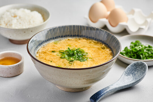 Japanese egg drop soup in ceramic bowl with scallion garnish, served beside steamed rice, soy sauce and raw eggs for fresh home style meal presentation, with soup, egg, broth, scallion detail