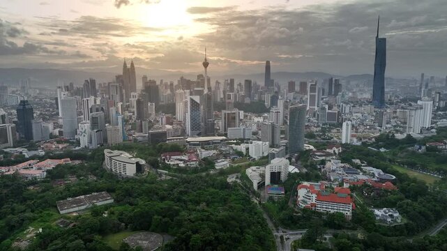 Aerial view of Kuala Lumpur city skyline at sunset featuring Merdeka 118, Petronas Twin Towers, and KL Tower with lush green forest in the foreground, Malaysia.