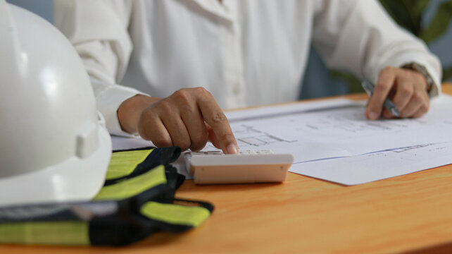 Construction engineer calculating project costs while reviewing architectural blueprints at a work desk with safety helmet and gloves. Professional planning, and construction management concept.