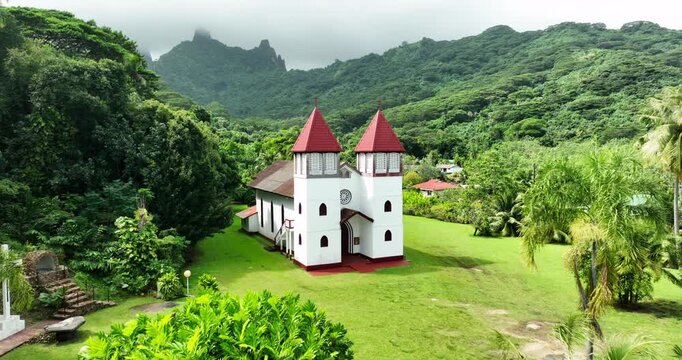 lisa Church's twin red-roofed spires rise above manicured lawn framed by palm trees, jagged volcanic peak looming behind dense tropical jungle valley in French Polynesia. Aerial drone view.
