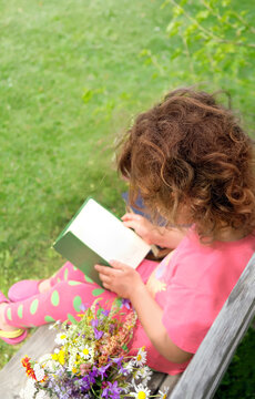 baby girl with curly hair sitting on wooden bench and reading book outdoor, natural background. Child studying in garden. education, reading hobby concept. unrecognizable person, without a face