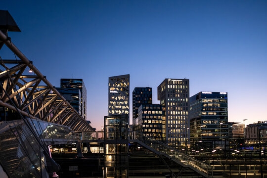 Beyond the dark framework, a steel bridge frames the skyline at twilight, while buildings and city lights reveal Oslo architecture beneath the evening sky of Norway after dusk