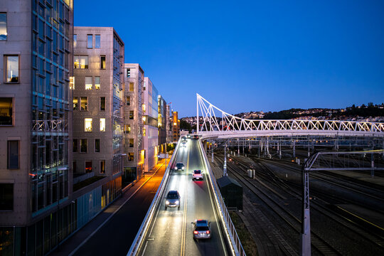 As evening settles into twilight over Norway, traffic follows the road beside a railway, while buildings reveal Oslo city lights through transportation infrastructure