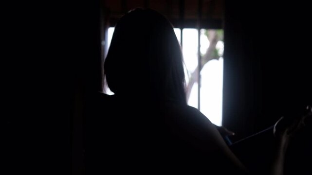 Silhouette of a young woman wrapping a towel around her shoulders looking out a bright window