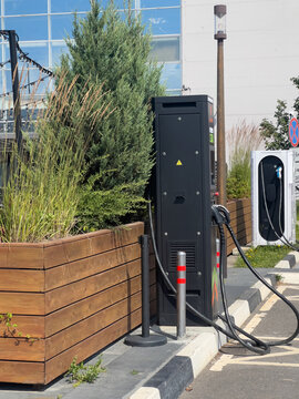 Electric vehicle charging station beside landscaped planter and lamp post in urban parking area with cables, connectors, glass building reflections and bright daylight