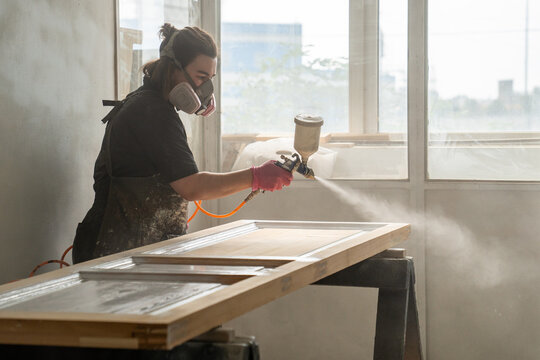 Restoration workshop process, craftsperson in paint booth finishing renovation of vintage wooden door, woman in respirator spraying paint from paint gun over surface, skills and knowledge at work.