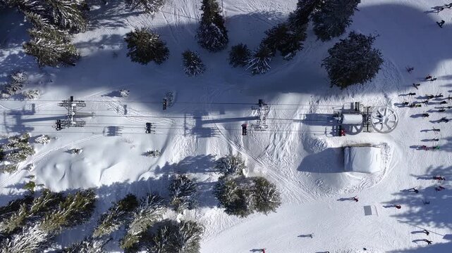 Aerial view over snowy mountain winter forest with chair lift at ski resort. Ski lift cable car top down drone shot. Winter landscape scene with snow covered trees. Outdoor tourism skiing snowboarding