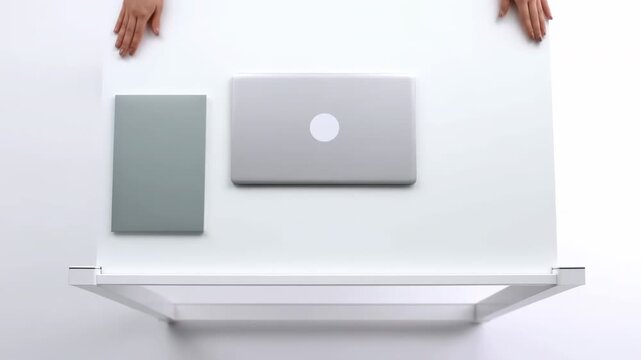 Overhead hands placing closed silver laptop beside gray notebook on white tabletop, clean workspace arrangement with minimal business styling, soft shadows, neutral tones, organized composition
