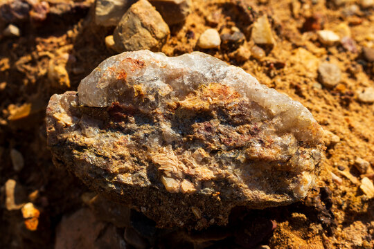 Close-up of a raw granite pegmatite specimen featuring coarse quartz and feldspar crystals resting on a rugged, sun-drenched gravel and sand quarry surface