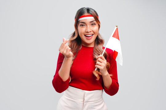 Smiling young Asian woman is holding a mini country flag while giving love finger gesture.