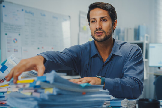 Overworked young Latin businessman reading document in large stack of paperwork files at office desk. Tax audit deadline, accounting stress, corporate burnout, bookkeeping, bureaucracy concept.