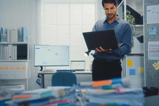 Thoughtful young Latin businessman stand with laptop analyzing data above paperwork stack. Hybrid work audit, digital transformation strategy, business corporate finance, executive decision.