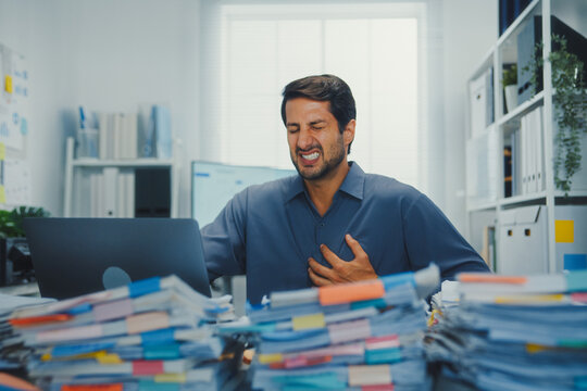 Stressed young Latin businessman having heart attack with severe chest pain at office desk. Cardiac arrest emergency, overworked burnout, medical stroke, work problem, health insurance crisis.