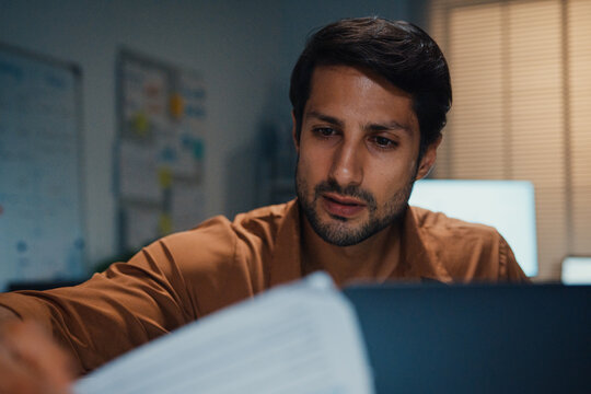 Close up overworked young Latin businessman working late on document in large stack of paper files at office night. Tax audit deadline, accounting stress, corporate burnout, urgent business concept.