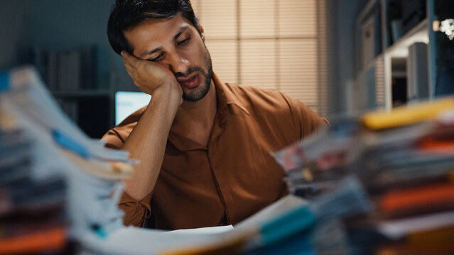 Overworked young Latin businessman waking from sleep nap to rush paperwork at desk in dark night office. Working late deadline burnout, heavy workload stress, and exhausted corporate problem concept.