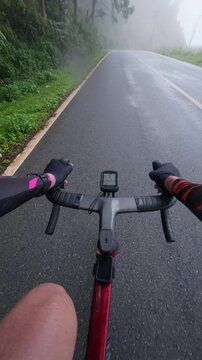 Vertical first-person POV footage of a road cyclist riding into dense mist on a wet mountain road surrounded by tropical forest. The reflective asphalt, soft fog, and peaceful atmosphere.