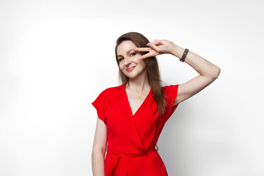 Young girl in red dress showing peace gesture near eye. Isolated on white background.