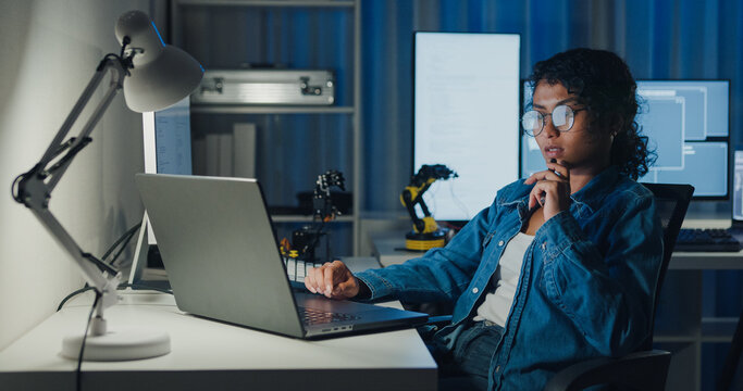 Young African American female programmer coding AI software on multiple screens in night office. Network security, cybersecurity, machine learning, working late, overtime, robotics automation concept.