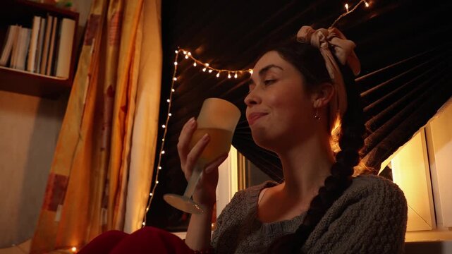 Low angle view of a young Caucasian woman relaxing at home in the evening with a glass of wine while enjoying peaceful solitude. Cozy lifestyle, self care, relaxation, and quiet evening concept.