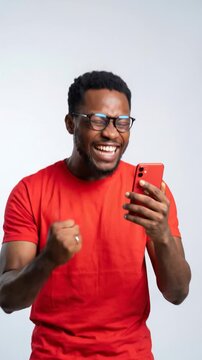 Happy young black man celebrating good news on smartphone while standing against a light studio background.