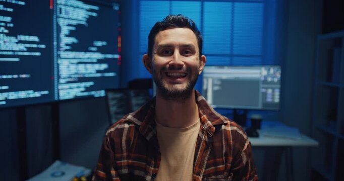 Portrait of confident Caucasian male programmer smiles looking at camera in office at night. Professional developer workspace, Secure computer network system, Advanced cybersecurity data concept.