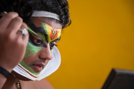 Kathakali dancer looking in the mirror and putting green colour paint on his face. 