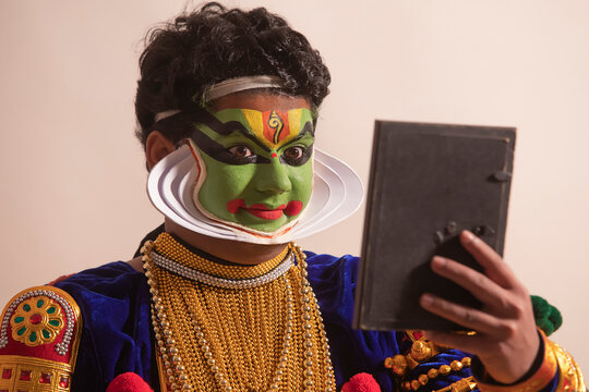 Kathakali dancer sitting in front of white background and checking his makeup in the mirror. 