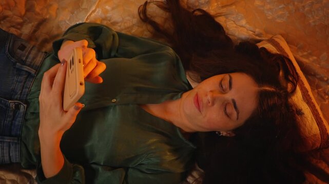 Top view of a young Caucasian woman with long hair lying on a bed and using a smartphone with an irritated and dissatisfied expression. Digital frustration, technology problems, and negative emotions 