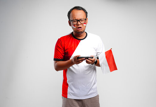 Crying young Asian man feeling sad looking at his smartphone while holding mini country flag.