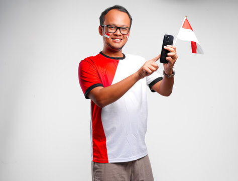 Happy young Indonesian man holding a mini flag while looking and using his cellphone.