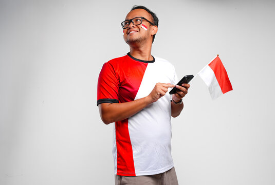 Happy young Indonesian man holding a mini flag while looking and using his cellphone.
