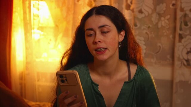 Portrait of a young Caucasian woman with long hair sitting on a bed and using a smartphone with a surprised expression. Digital communication, online news, technology, and modern lifestyle concept
