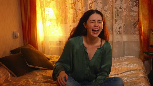 Mid shot of a cute young woman laughing happily while sitting on a bed in a cozy evening room. Joyful emotions, relaxation, comfort, and positive home lifestyle concept.