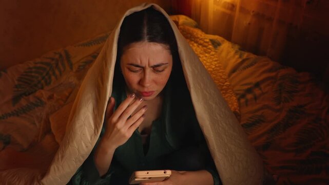 Top view close up of a frightened woman wrapped in a blanket using a smartphone while discovering bad news online. Shock, fear, anxiety, and emotional reaction concept.