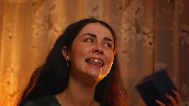 Portrait of a joyful young Caucasian woman with braces excitedly opening a gift box and discovering a surprise in a cozy evening room. Happiness, celebration, confidence, and positive emotional reacti