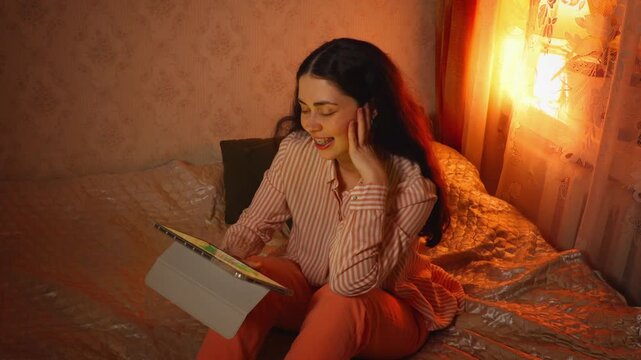Top view of a relaxed young Caucasian woman watching videos on a tablet while sitting on a bed. Cozy home atmosphere, digital entertainment, leisure, and evening relaxation concept
