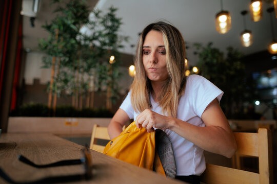 Frustrated Woman Searching For Something In Her Backpack At Restaurant. Stressed lady looks inside open bag on table trying to find lost item