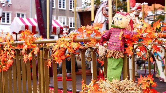 Scarecrow doll displayed with pumpkins and harvest decorations in outdoor urban setting near carousel. Seasonal arrangement features rustic autumn elements, colorful foliage and festive ornaments