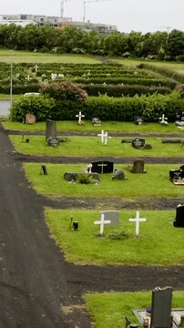 An aerial view of a cemetery with green grass, headstones, and white crosses. In the background, a new construction site with cranes is visible beyond the trees.