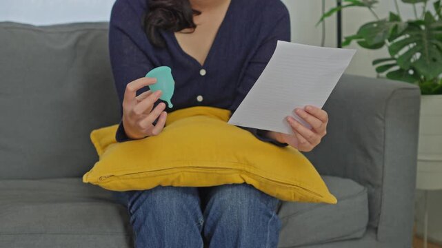 Woman sitting reading paper instructions while holding reusable silicone menstrual cup on sofa