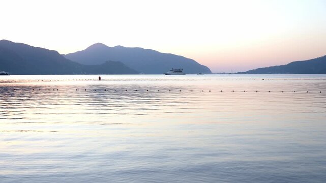 Scenic waterfront in Marmaris, Turkey during blue hour at sunrise or sunset. The coastal promenade overlooks calm sea framed by mountain silhouettes in the distance. Small boat floats gently on waves