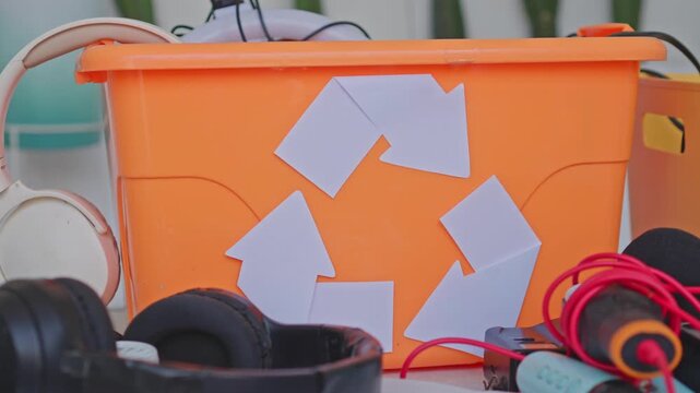 Orange container with paper recycling symbol filled with discarded electronics and headphones