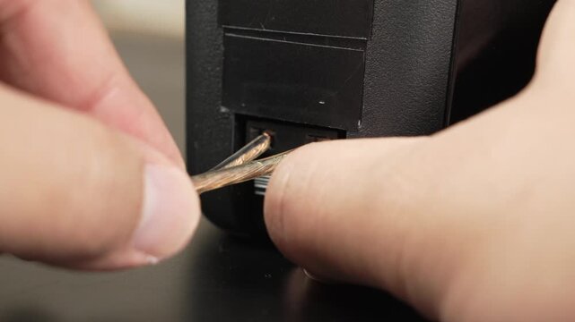 Hands attach bare speaker wire to the spring clip terminals on the back of a black bookshelf speaker on a desk, useful for tutorials, product demos, or home o guides.