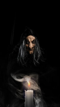 A creepy old witch with a wrinkled face and dark hood glares from the shadows behind a smoking, burning white candle. Concept of Halloween