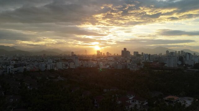 Da nang city skyline at sunset, orange sky with clouds reflecting golden light, urban scene contrasting with distant mountains and dark foreground trees