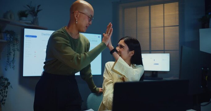 Young Asian businesswomen team giving high five at office desk at night. Corporate teamwork success celebration, professional colleague partner support, happy employee project achievement win concept.
