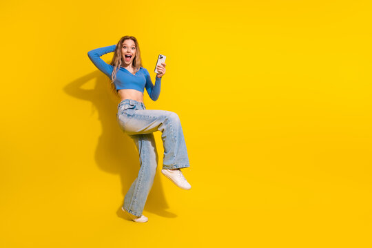 Young woman joyfully holding smartphone against yellow background expressing surprise and excitement