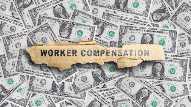 A piece of torn cardboard with the text WORKER COMPENSATION sits on top of a background of many United States one-dollar bills, representing financial benefits. Concept of Compensation