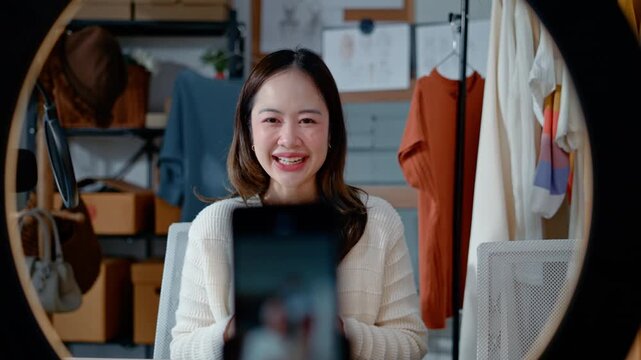 Young Asian woman fashion entrepreneur live streaming selling clothes online with smartphone ring light and laptop in home studio, representing small business owner and professional women.